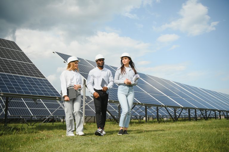 Group of multi ethnic people and safety helmets staring at solar farm and looking on drawings. Two engineers and technician examining plan of photovoltaic construction outdoors.